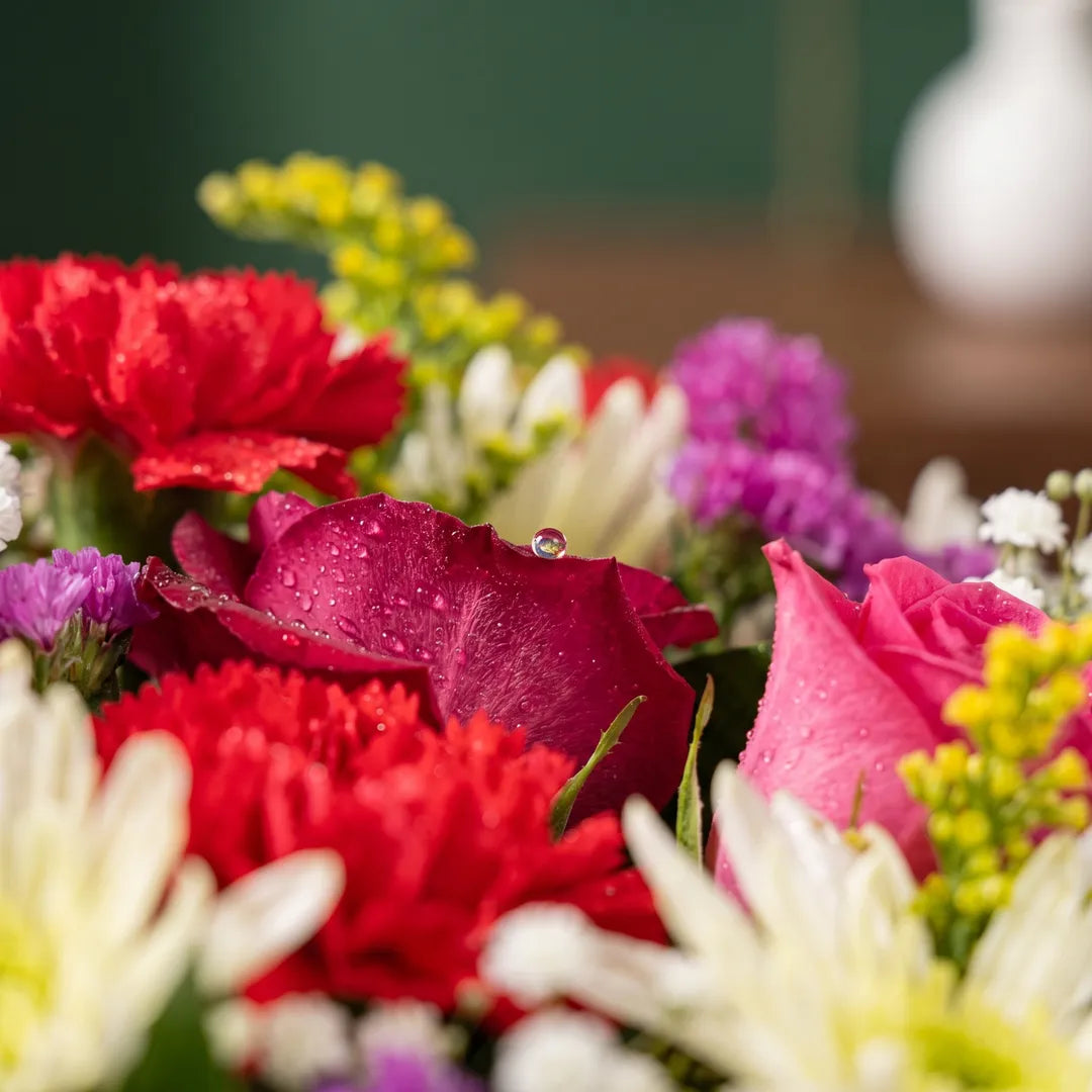 A close-up bouquet of mixed flowers including red roses, pink carnations, purple blossoms, white chrysanthemums, and yellow sprigs with water droplets on the petals.