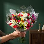 A vibrant mixed flower bouquet wrapped in clear polka-dot tissue paper, held by a person's hands against a dark green background with a wooden side table.