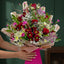 A large, vibrant floral bouquet held by hands against a dark green background. The bouquet is a rich mix of red roses, white daisies, pink lilies, purple alstroemeria, and green foliage, wrapped in patterned brown paper and a bright pink base box.