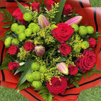 A vibrant floral bouquet featuring red roses, bright green chrysanthemums, pink lily buds, and green ferns, wrapped in red paper with black stripes and placed on a green grass-like surface.