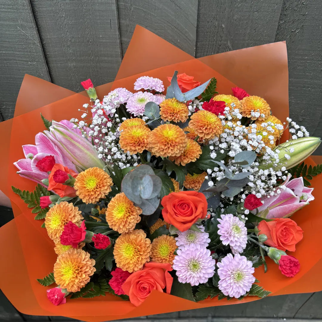 A vibrant mixed flower bouquet wrapped in bright orange paper, featuring orange and yellow chrysanthemums, pink and orange roses, purple chrysanthemums, baby's breath, and lily buds, set against a dark gray wooden background.