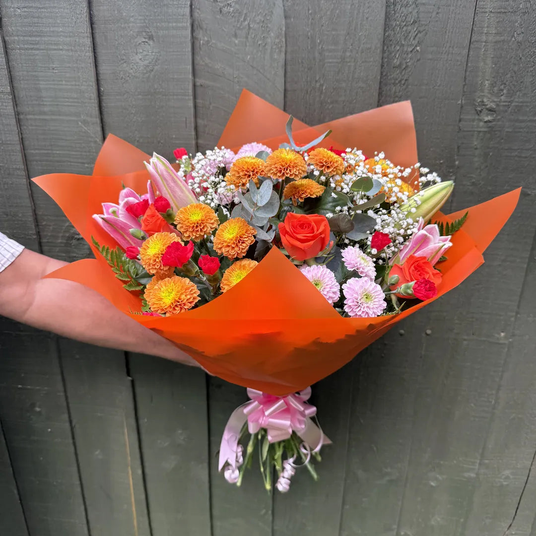 Person holding a large mixed bouquet of orange chrysanthemums, coral roses, pink lilies, cerise carnations and gypsophila in orange wrap with pink ribbon -- Blossom Surprise Northampton