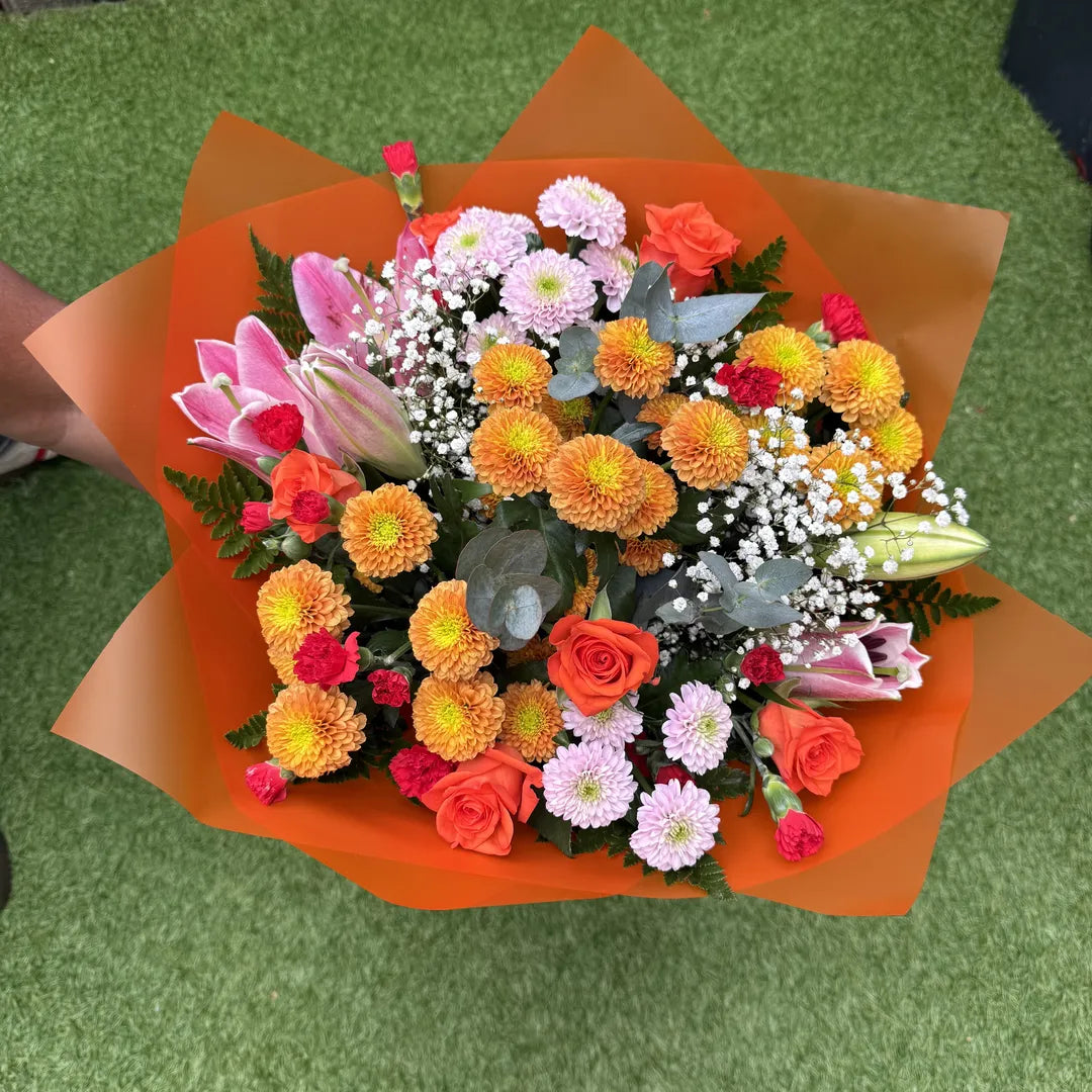 A vibrant mixed flower bouquet wrapped in bright orange paper, featuring orange roses, pink lilies, yellow and orange chrysanthemums, and white baby's breath, set against a green artificial turf background.