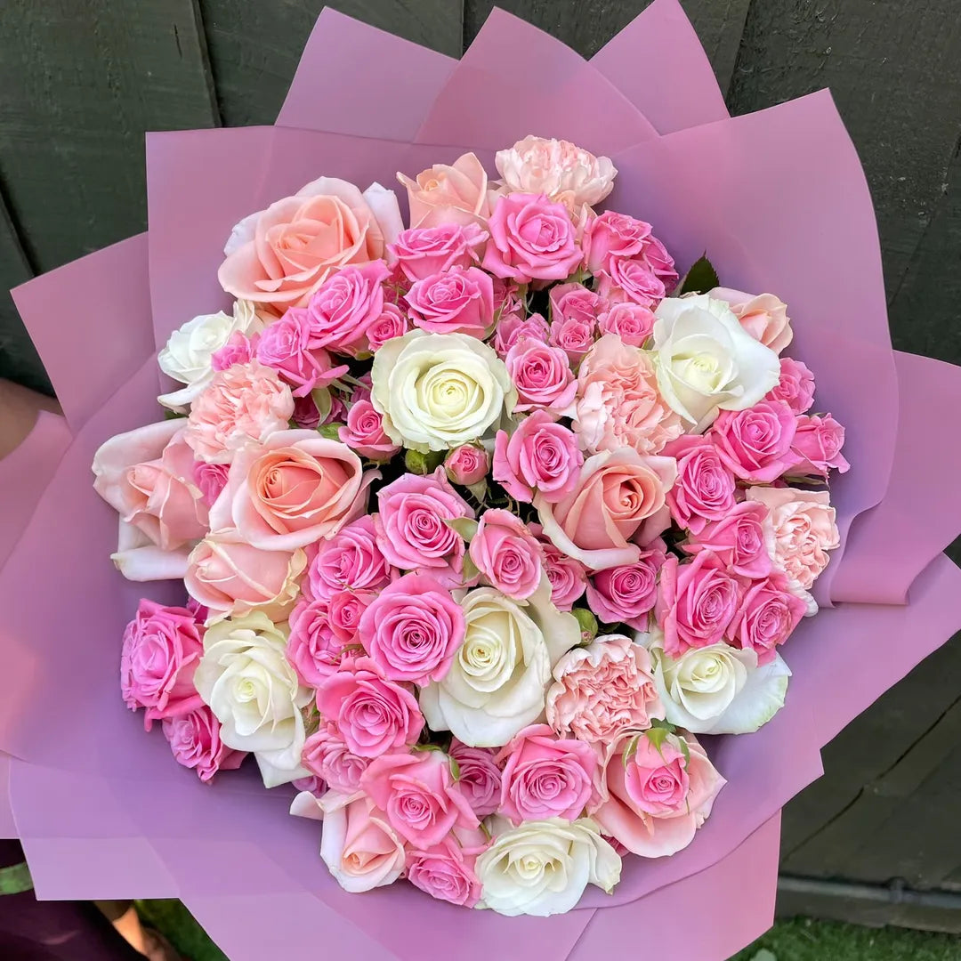 A round bouquet of pink and white roses wrapped in light purple paper, photographed outdoors against a dark wooden fence.