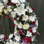 Detail of open ring funeral wreath showing fully open white lily, white chrysanthemums, red roses and soft purple statice -- Blossom Surprise Northampton
