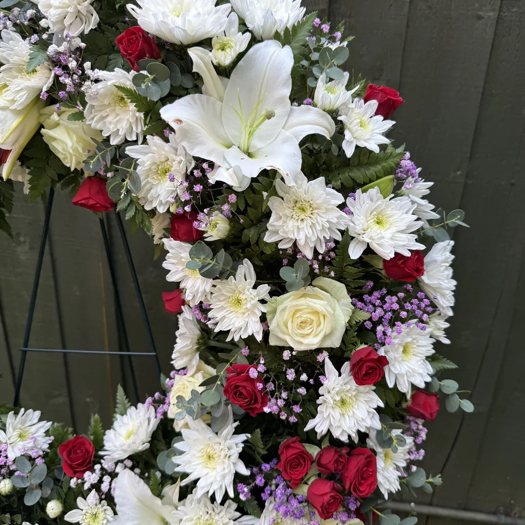Detail of open ring funeral wreath showing fully open white lily, white chrysanthemums, red roses and soft purple statice -- Blossom Surprise Northampton