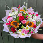 Hand holding a mixed bouquet of sunflowers, red and pink roses, carnations, white chrysanthemums and gypsophila with grass blades in blush marble wrap -- Blossom Surprise Northampton