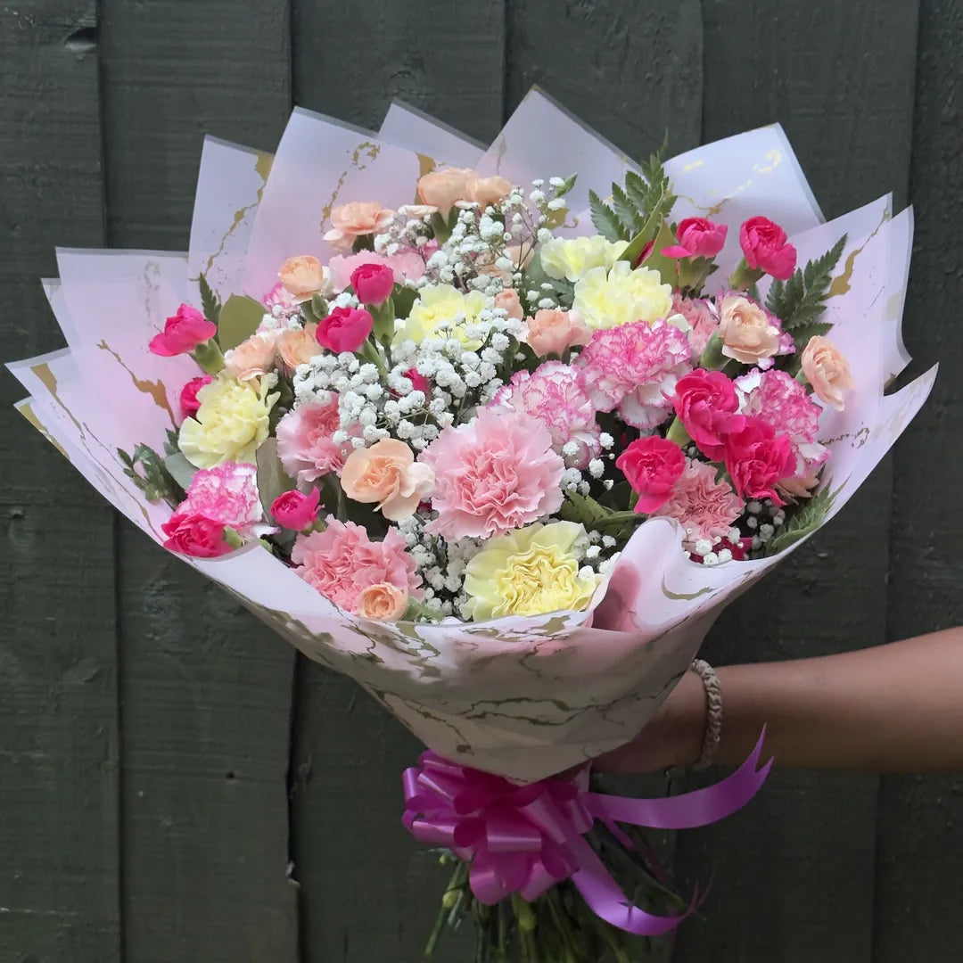 Hand holding a full pastel carnation bouquet with bicolour frilled carnations, gypsophila and fern in blush wrap -- Blossom Surprise Northampton